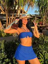 Woman in a blue bikini and skirt posing outdoors with a hat and sunglasses.