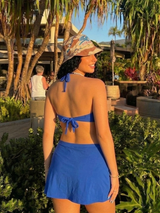 Woman in a blue bikini and hat standing outdoors with greenery and people in the background
