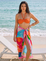Woman in an orange bikini top and colorful sarong on a beach.