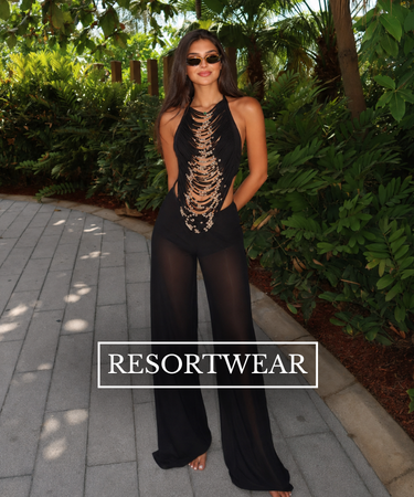 A woman wearing a black sheer sleeveless jumpsuit with gold chain detailing on the bodice, standing outdoors on a paved walkway surrounded by green foliage. A 'RESORTWEAR' brand label is overlaid at the bottom.