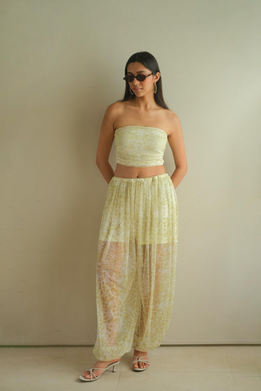 A woman modeling a pale yellow lace two-piece set consisting of a strapless crop top and matching loose harem pants, paired with clear high-heeled sandals and sunglasses, standing against a plain beige wall.