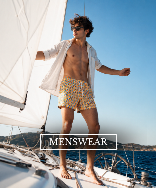 A man stands on a sailboat wearing patterned swim trunks and an open white shirt, with blue sea and sky in the background and a 'MENSWEAR' text overlay.