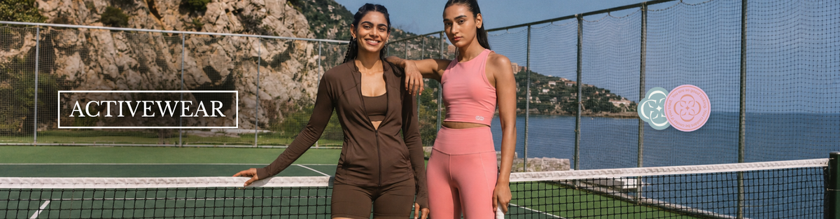 Two women posing on a tennis court wearing activewear from ActiveWear, with a rocky coastline and sea in the background. One woman wears a dark brown two-piece set and the other wears a pink two-piece set.