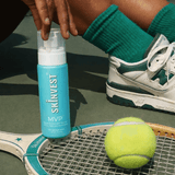 Person holding a Skinvest MVP bottle on a tennis court with a tennis ball and racket.
