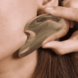 Person using a bronze Gua Sha tool on their neck with 'Skinsvest' branding.