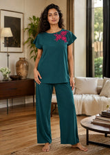 Woman wearing teal pajama set with floral embroidery in a living room.