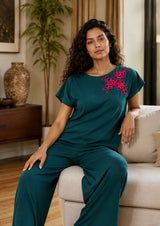 Woman in teal outfit with pink floral embroidery sitting on a couch in a living room.
