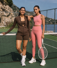 A woman posing on a green tennis court wearing a white athletic crop top and matching white tennis skirt by ActiveWear, holding a pink racket handle and a tennis ball, with a tennis racket behind her.