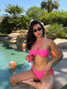 Woman in a pink bikini holding a can by a pool with beach balls and palm trees in the background