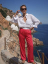 Woman in white blouse and red pants standing on a scenic coastal path with blue sea and white domes in the background.