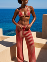 Woman in a red and white striped bikini and pants standing by the ocean.