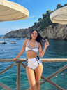 Woman in a white bikini with blue accents standing by a railing with a scenic ocean view.
