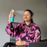 Woman in a pink and black patterned blouse holding a blue spray bottle in an office setting.