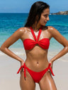 Woman in a red bikini standing on a beach with clear blue water and sky.